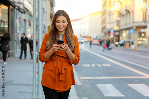 Portrait of beautiful smiling woman walking in city street texting on mobile phone with blurred background