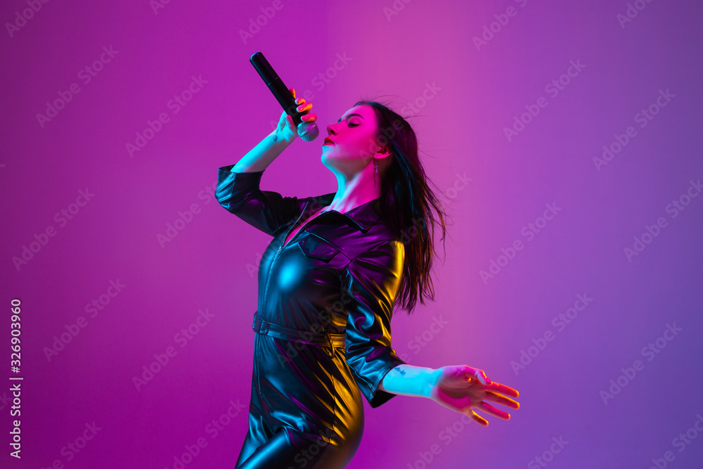 Caucasian female singer portrait isolated on purple studio background in neon light. Beautiful female model in black wear with microphone. Concept of human emotions, facial expression, ad, music, art.
