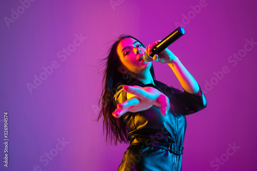 Caucasian female singer portrait isolated on purple studio background in neon light. Beautiful female model in black wear with microphone. Concept of human emotions, facial expression, ad, music, art. photo