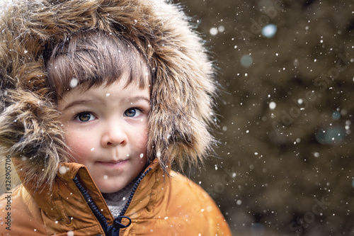 Close up portrait of cute little toddler boy with rosy cheeks in furry hood at snowfall outdoors looking at camera. Winter lifestyle. Snowy weather