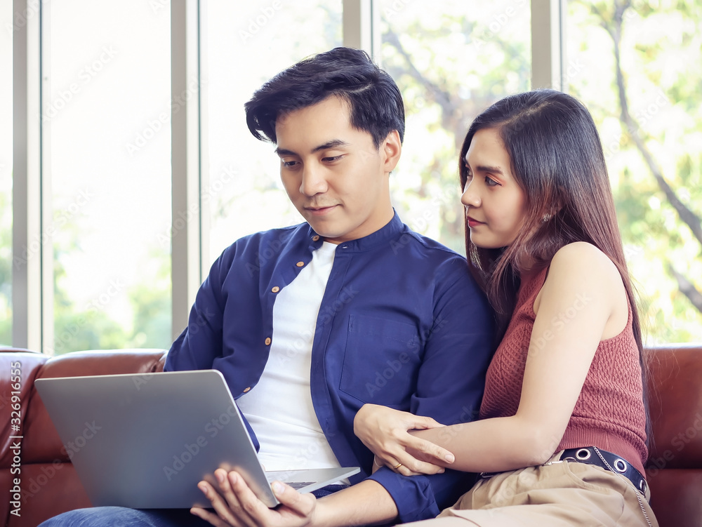Young Asian couple sitting close together on couch in living room , a man using computer while woman holding his arm , smiling and looking at his face Young Asian couple sitting close together on couch in living room , a man using computer while woman holding his arm , smiling and looking at his face