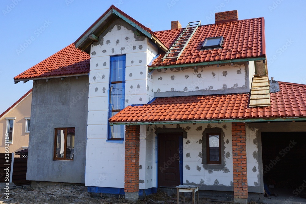 large unfinished private house with white insulation on a gray wall under a red tile roof against a blue sky on a sunny day