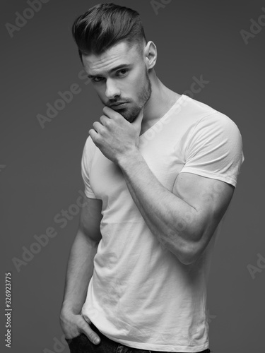  Monochrome portrait of a young man with a beard on a gray background. A muscular man in a t-shirt. Studio portrait. Fitness, fashion and beauty