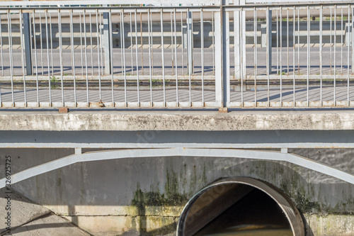 A concrete small bridge with a pedestrian path, asphalt road surface, gray metal fences and with a reinforced concrete slab and a metal pipe under it, along which a stream flows