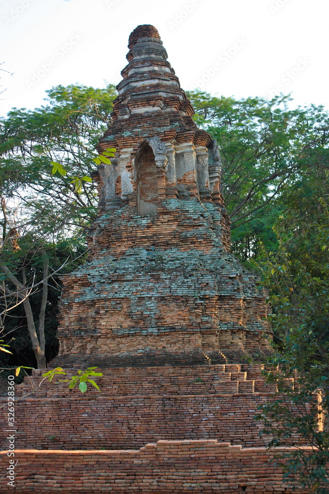 Ancient pagoda being ruined and left only red brick architecture with some old time concrete. Tall green tree on background and white sky. The historical landscape of Chiang Mai town.