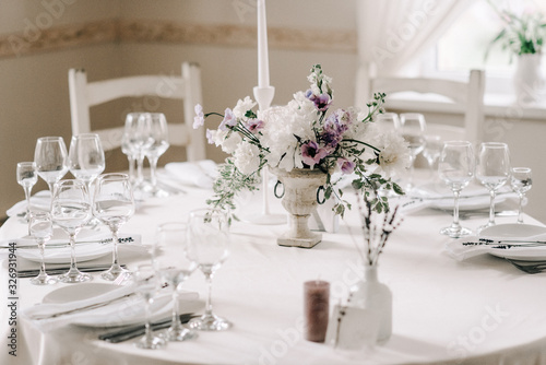 Wedding banquet table setting. Plates, glasses, cutlery and flower arrangement on a white round table. Round table with a white tablecloth. Plate with a gray cloth napkin.