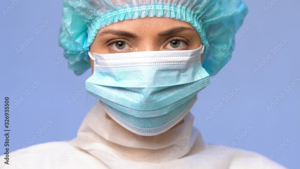 young woman doctor with a medical cap on her head puts on a medical face mask, looks seriously at the camera, close-up portrait on a blue background. copy space. 2019 Novel Coronavirus concept young woman doctor with a medical cap on her head puts on a medical face mask, looks seriously at the camera, close-up portrait on a blue background. copy space. 2019 Novel Coronavirus concept