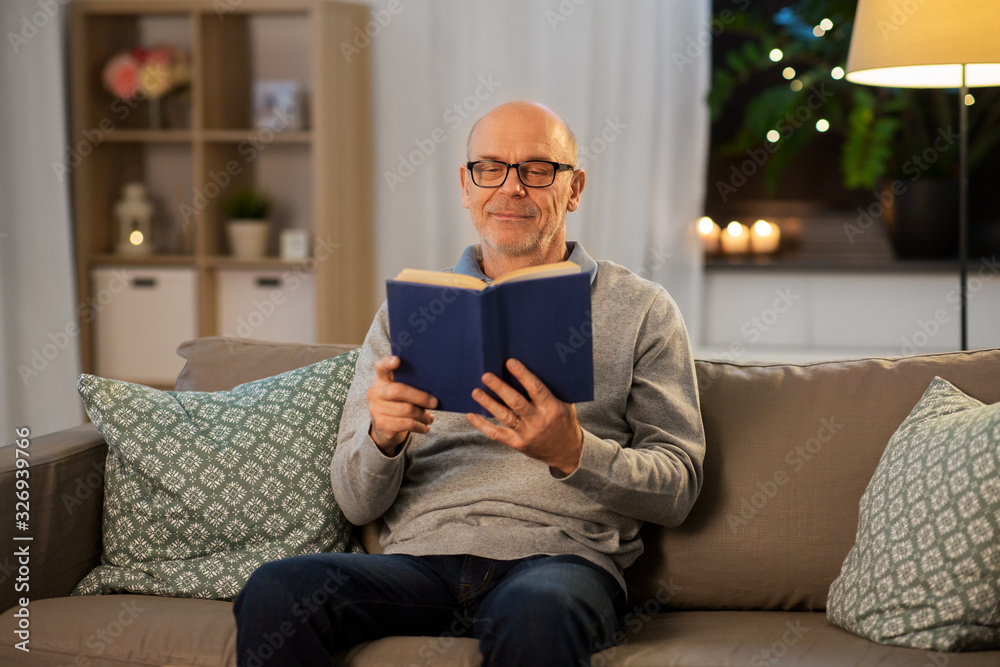 old age, leisure and people concept - happy bald senior man sitting on sofa and reading book at home in evening old age, leisure and people concept - happy bald senior man sitting on sofa and reading book at home in evening
