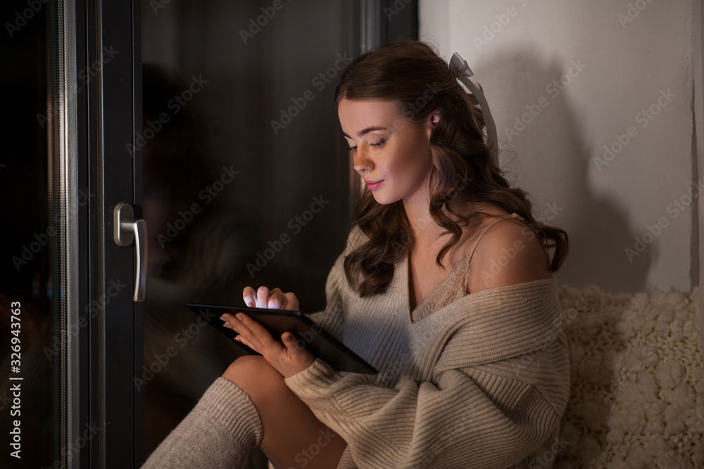 winter, comfort and people concept - young woman in pullover with tablet pc computer sitting at window at home at night winter, comfort and people concept - young woman in pullover with tablet pc computer sitting at window at home at night