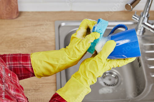 Hands with sponge wash the cup under water, housewife in protective gloves manually washing blue mug. Hand cleaning