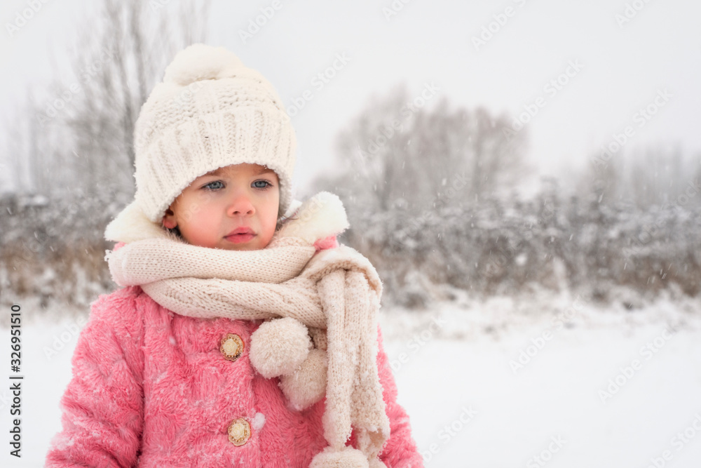little girl in a purple coat, white hat and a large scarf is sad in winter during a snowfall