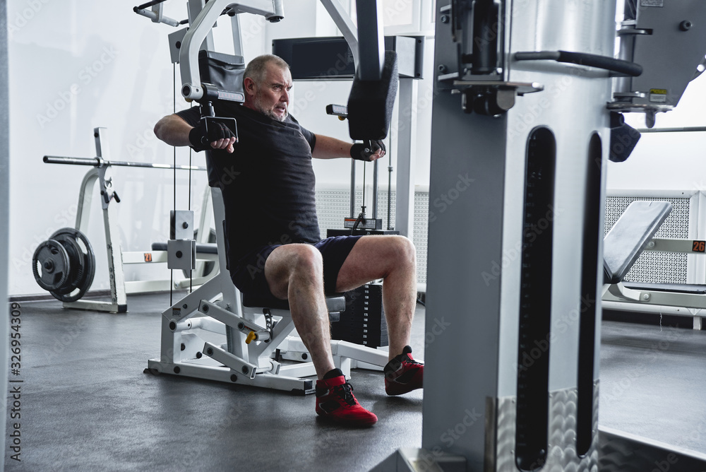 Adult gray-haired man trains on fitness equipment in the gym, pumps legs and arms muscles, loses weight. Concept of healthy lifestyle in old age Adult gray-haired man trains on fitness equipment in the gym, pumps legs and arms muscles, loses weight. Concept of healthy lifestyle in old age