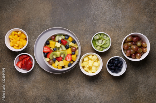 Fruit salad in bowl with ingredients. Multi-colored ripe fruits and berries. Pineapple, mango, grape, strawberry, blueberry and kiwi