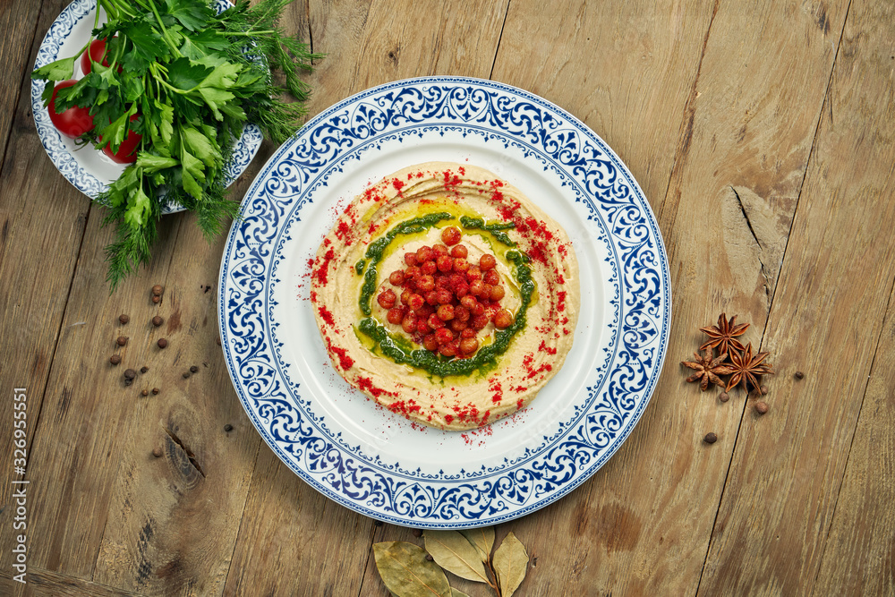 Hummus with red spices, chickpeas and herbs on a ceramic plate on a wooden background. Arabic or Israeli cuisine. Close up, horizontal. Snack Hummus with red spices, chickpeas and herbs on a ceramic plate on a wooden background. Arabic or Israeli cuisine. Close up, horizontal. Snack