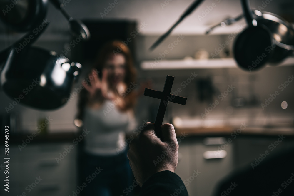 Selective focus of female demon with levitating cookware and exorcist with cross in kitchen
