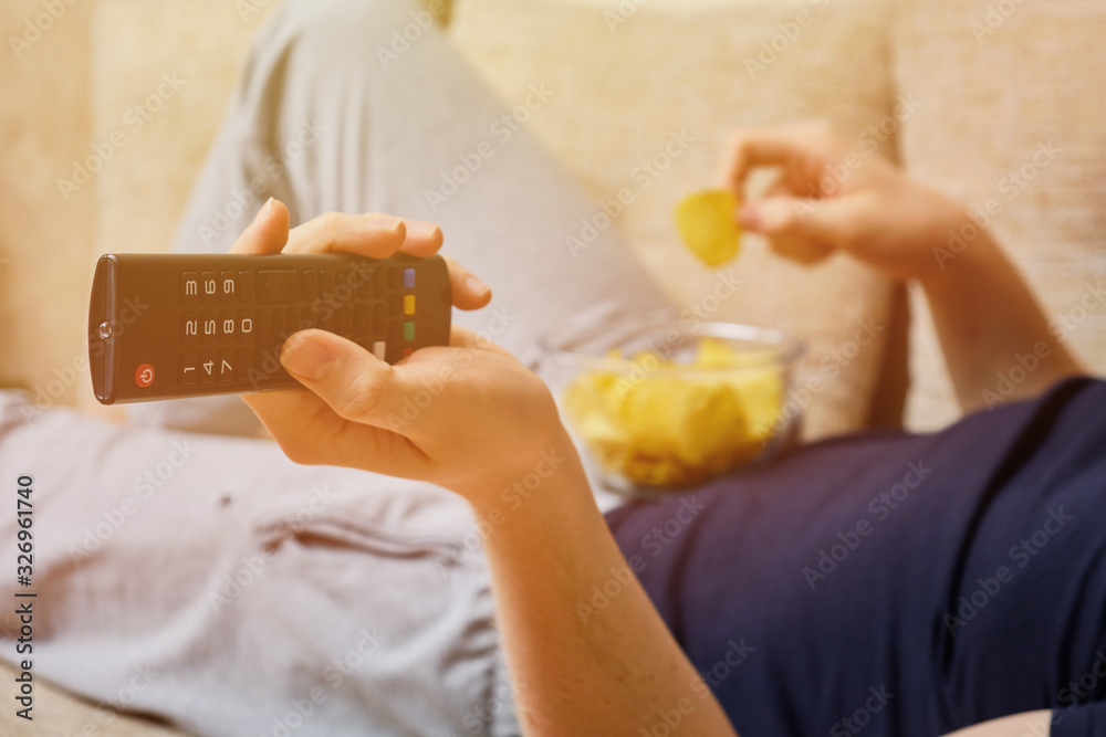 A man lies and rests on a sofa with a remote control from the TV, changing channels, eating potato chips from glass dishes.