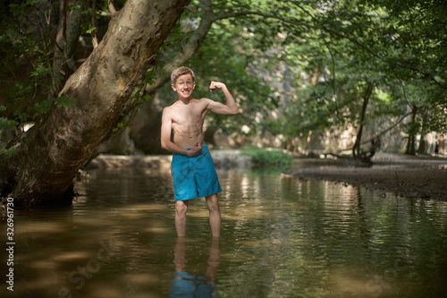 Smiling happy teenage boy having fun by posing like bodybuilder in shallow water in sunny forest in mediterranean country in summer time  relaxed mood fun smile happiness
