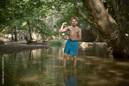 Smiling teenage boy posing like bodybuilder in shallow water in forest in mediterranean country in summer time, sunny relaxed mood fun smile happiness
