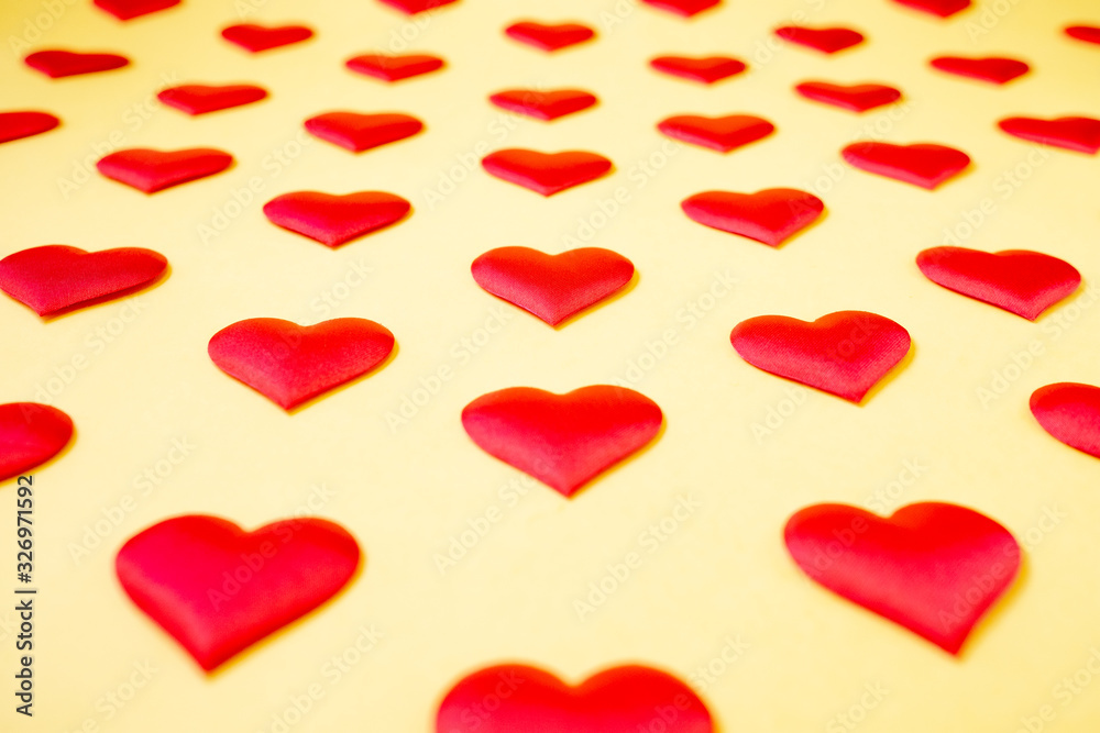 A lot of identical red silk hearts lying staggered on a yellow background. Symbol of love, tenderness and passion