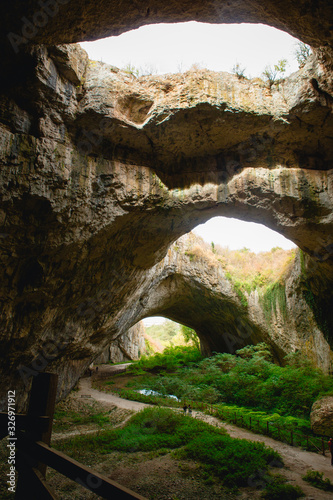 Fototapeta Naklejka Na Ścianę i Meble -  High arches of a huge stone cave with round holes at the top, a tourist road with a fence inside the cave. Bulgaria's natural attraction-Devetashka cave