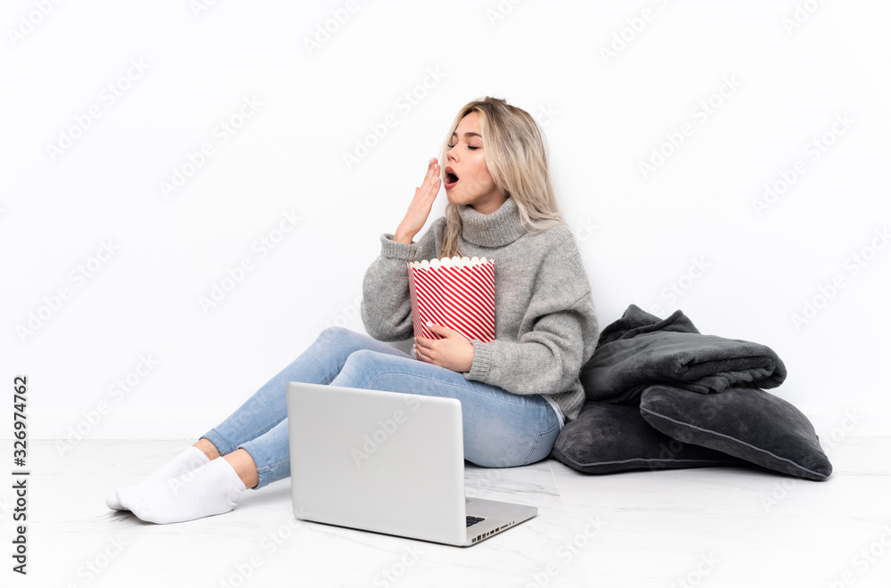 Teenager blonde girl eating popcorn while watching a movie on the laptop yawning and covering wide open mouth with hand Teenager blonde girl eating popcorn while watching a movie on the laptop yawning and covering wide open mouth with hand
