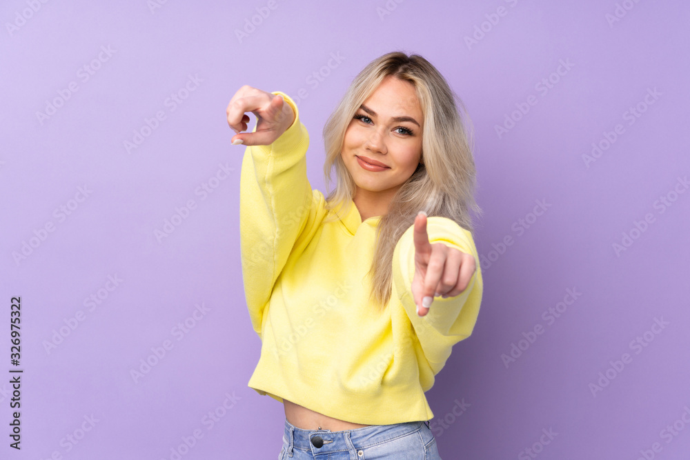 Teenager girl wearing a yellow sweatshirt over isolated purple background points finger at you while smiling Teenager girl wearing a yellow sweatshirt over isolated purple background points finger at you while smiling