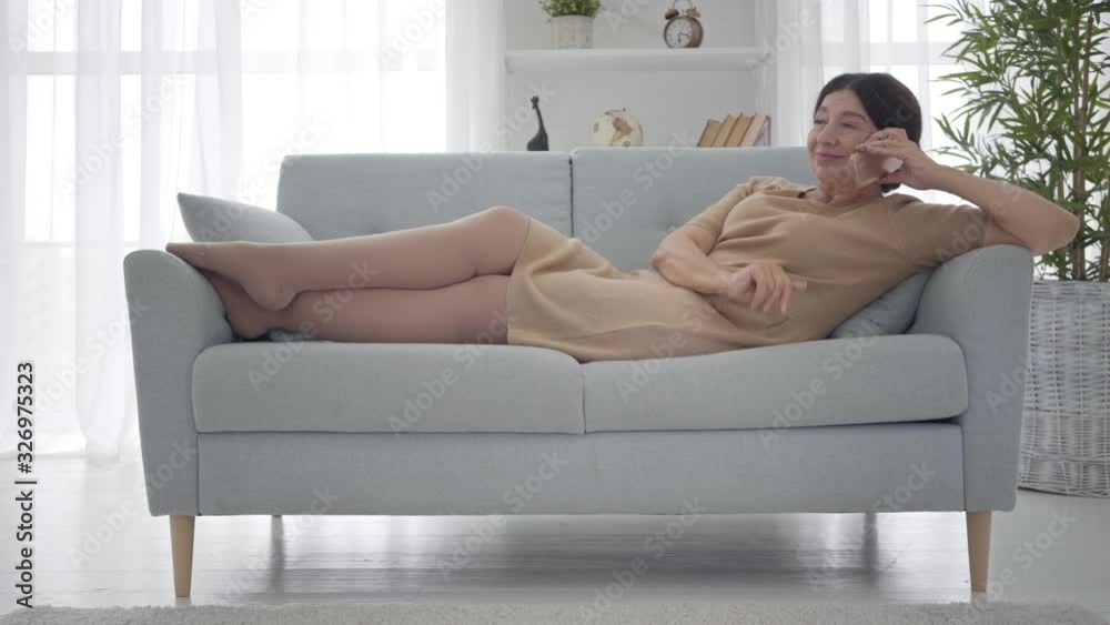 Relaxed Caucasian senior woman in elegant beige dress lying on sofa and talking on the phone. Happy middle-aged female retiree spending free time at home. Lifestyle, leisure, tranquility. Wide shot.