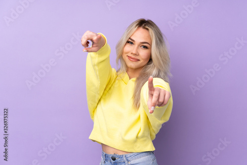 Teenager girl wearing a yellow sweatshirt over isolated purple background points finger at you while smiling