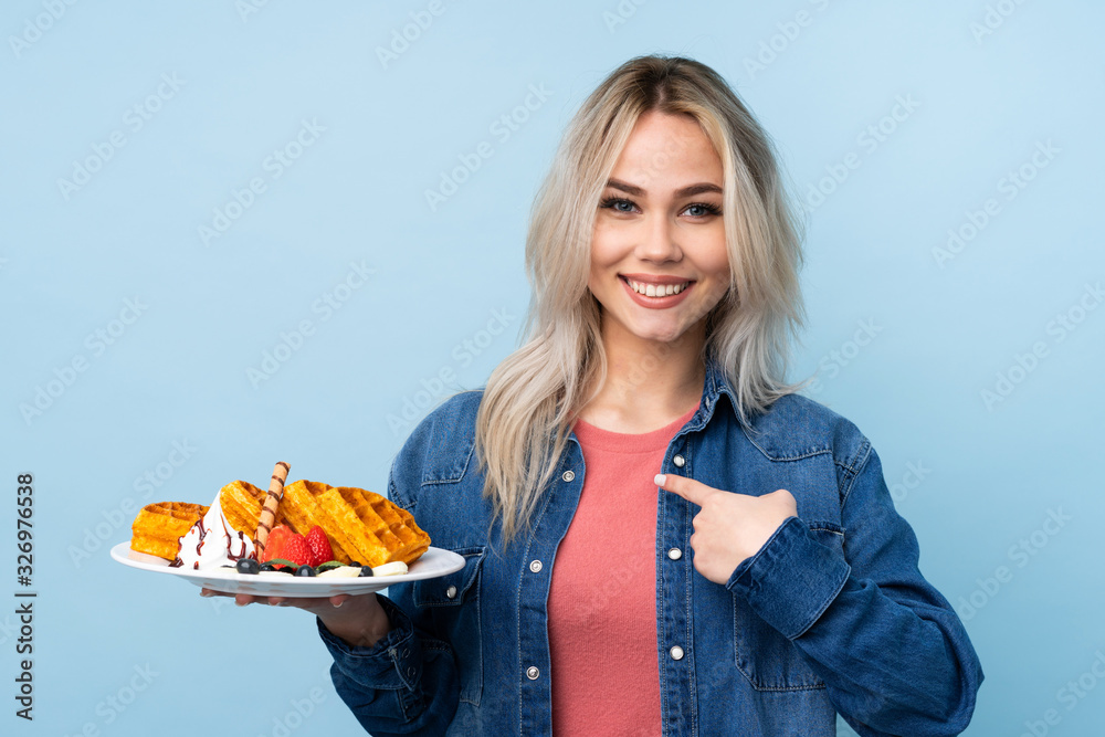 Teenager girl holding waffles over isolated blue background with surprise facial expression Teenager girl holding waffles over isolated blue background with surprise facial expression