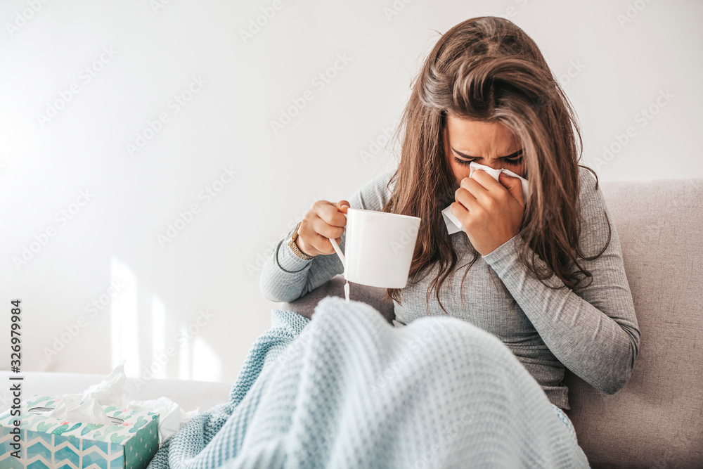 Sick woman blowing her nose. Sick young woman at home on the sofa with a cold, she is covering with a blanket and blowing her nose. Woman drinking hot tea and feeling comfortable Sick woman blowing her nose. Sick young woman at home on the sofa with a cold, she is covering with a blanket and blowing her nose. Woman drinking hot tea and feeling comfortable
