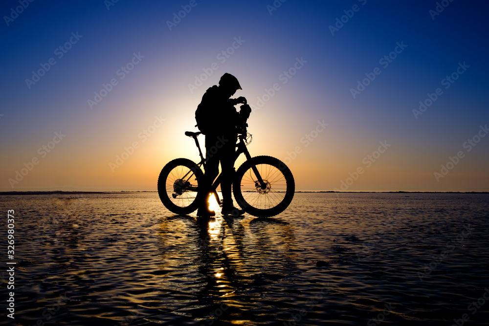 Fototapeta premium Silhouette of bicyclist standing on the clear ice surface of frozen lake in morning backlight