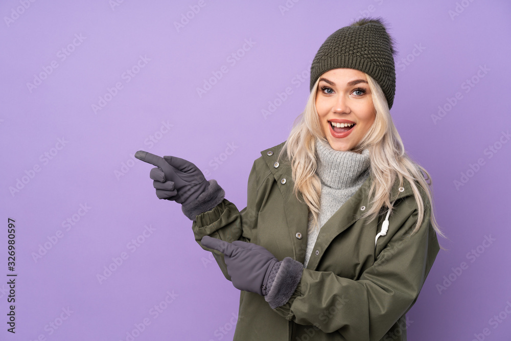 Teenager blonde girl with winter hat over isolated purple background surprised and pointing side Teenager blonde girl with winter hat over isolated purple background surprised and pointing side