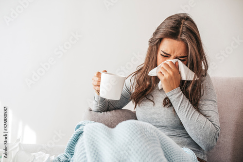 Sick woman covered with a blanket lying in bed with high fever and a flu, blowing her nose. Pills and glass of water on the table. Woman sneezing in a tissue in the living room photo