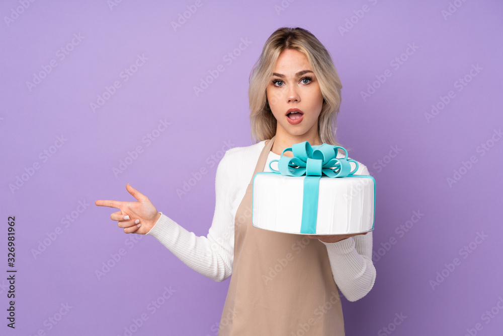 Young pastry chef holding a big cake over isolated purple background surprised and pointing finger to the side Young pastry chef holding a big cake over isolated purple background surprised and pointing finger to the side