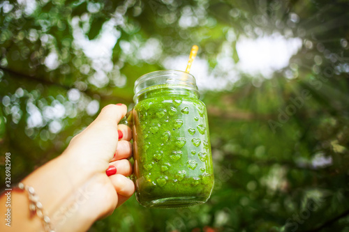 woman's hand holding jar with green cold-pressed juice, vegetable garden background. Healthy eating, detoxing, juicing, body cleancing concept photo