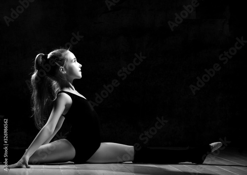 Young smiling girl gymnast with long hair in black sport body and uppers sitting in twine sideways over dark background