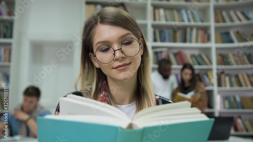 Pretty calm concentrated young woman in glasses reading book in the university library photo
