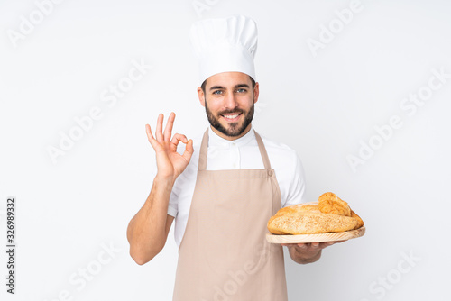 Male baker holding a table with several breads isolated on white background showing an ok sign with fingers