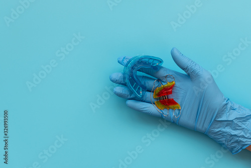 Hand of medic wearing blue latex glove holding myofunctional trainer and retainer orthodontic appliance on blue background. Teeth correction concept photo