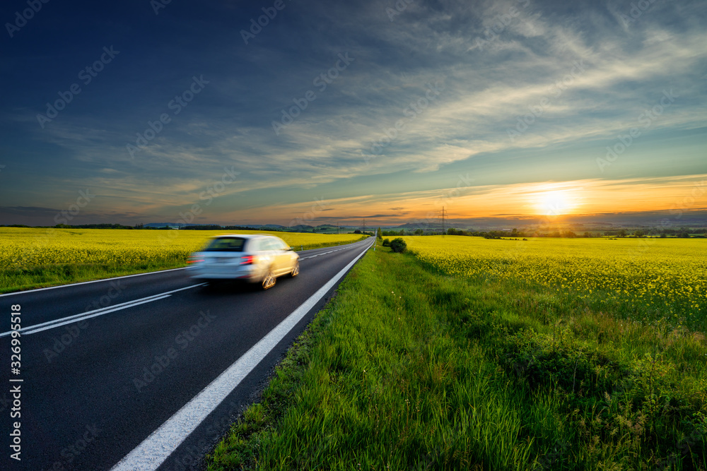 Motion blurred white car driving on the empty asphalt road between the yellow flowering rapeseed fields in the rural landscape at sunset