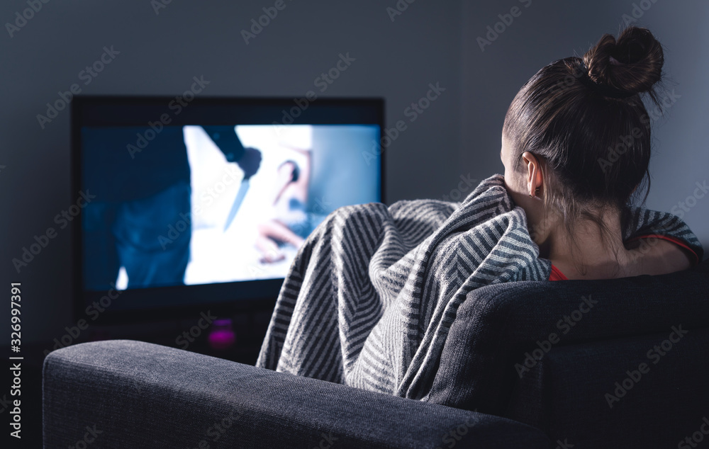 Scary horror movie on tv. Scared woman watching stream service hiding under  blanket on couch at