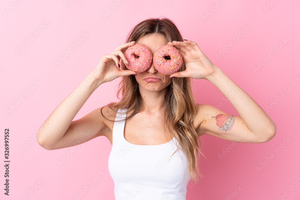 Young woman over isolated pink background holding donuts in eyes with sad expression