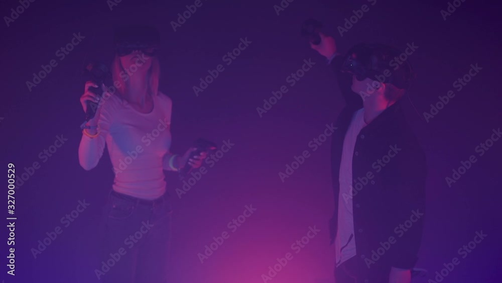 Young Man and Women Using Virtual Reality Glasses Holding Joysticks Controllers, Moving Hands, Loking Around Standing in the Room with Neon Lighting Colors Background. Young Man and Women Using Virtual Reality Glasses Holding Joysticks Controllers, Moving Hands, Loking Around Standing in the Room with Neon Lighting Colors Background.
