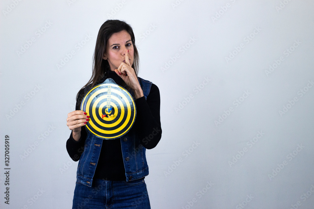 Portrait of attractive caucasian middle age woman with dart board holding index finger near his lips, silence, isolated on gray background studio shot, black sweater, denim jacket, jeans, dark air. Pl