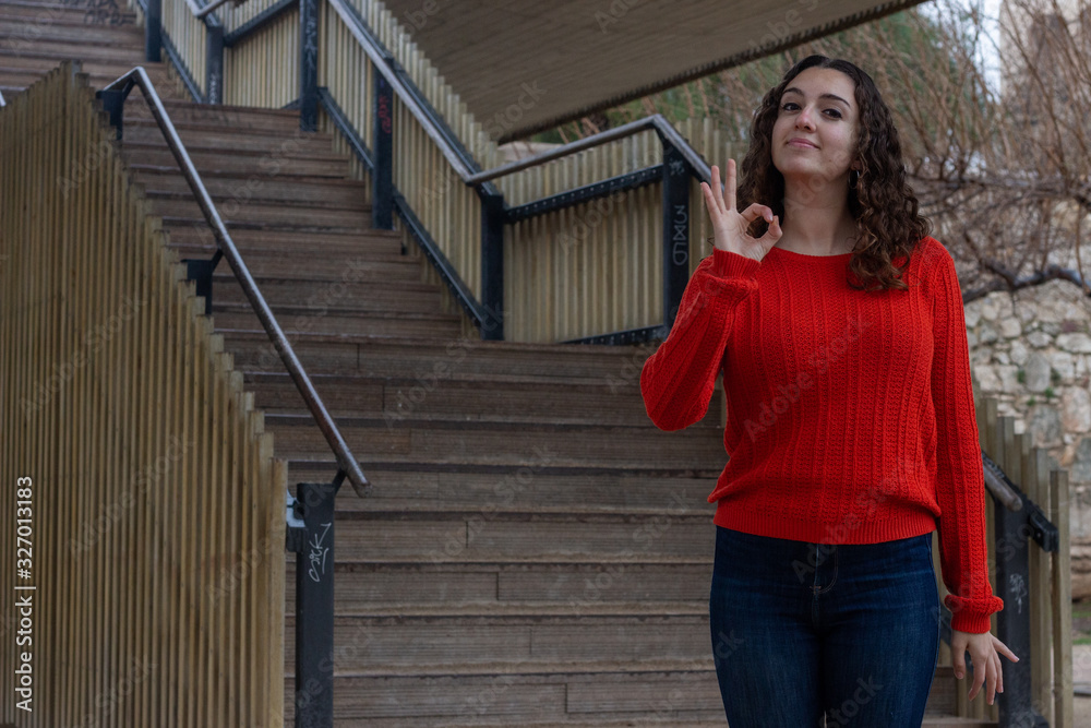 Portrait of positive caucasian young woman model showing ok sign with hand and fingers, has satisfied facial expression, in the park, orange sweater and jeans, long curly hair. Place for your text in