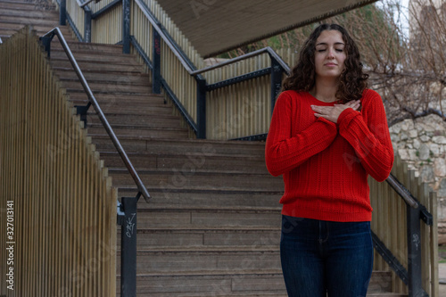 Portrait of caucasian young woman with closed eyes, keeps hands on chest, in the park, orange sweater and jeans, long curly hair. Place for your text in copy space.