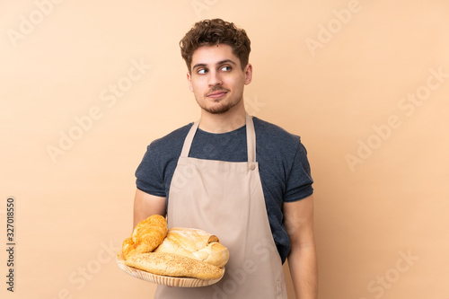 Male baker holding a table with several breads isolated on beige background standing and looking to the side