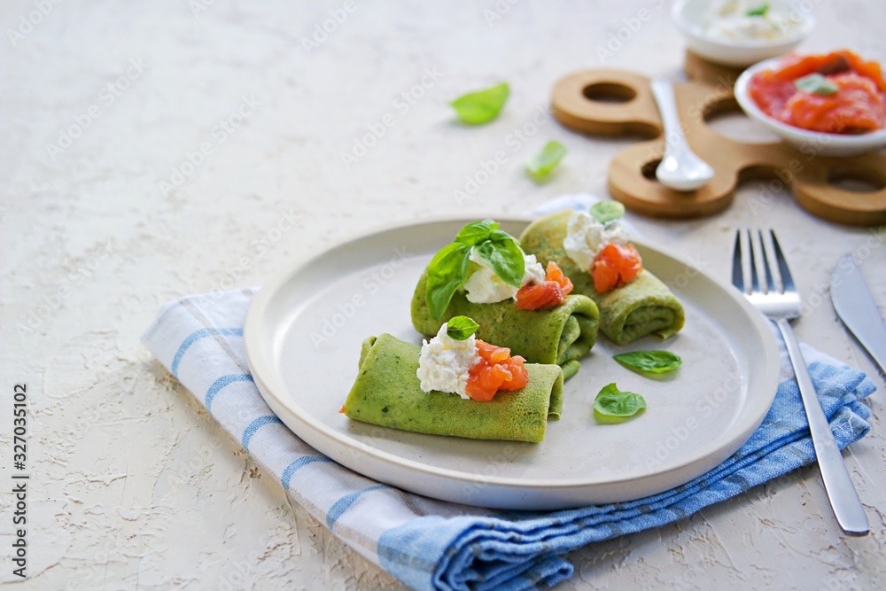 Spinach pancakes rolled up on a white plate on a light concrete background. Served with salted salmon and soft cheese. Spinach recipes. Copyspace. Spinach pancakes rolled up on a white plate on a light concrete background. Served with salted salmon and soft cheese. Spinach recipes. Copyspace.