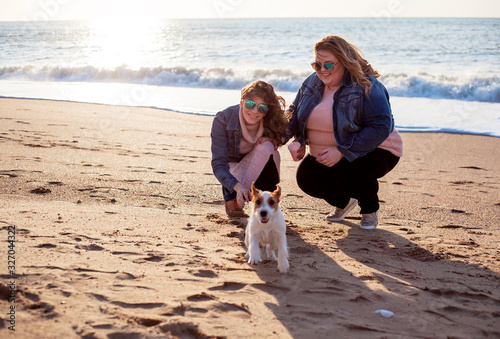 Two freinds and dog walking on the spring beach. Fat woman with strong friend playing with pet. Overweight woman dressed jeans jacket and pink sweater. people enjoing the nature and evening sun