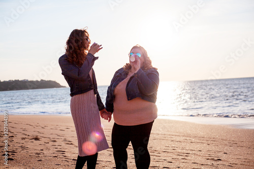 Two freinds , plus size with  thin  girls walking on the spring beach. Fat woman with strong friend drinking tea. Overweight woman dressed jeans jacket and pink sweater.  photo